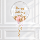 A clear round birthday balloon with gold script reading 'Happy Birthday Emily,' attached to pink and gold mini balloons, a white ribbon, and a gold curling ribbon against a white paneled wall.