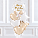 A birthday balloon bouquet with a large clear round balloon reading 'Happy Birthday Roisin' in gold script, plus metallic heart, star, and small round balloons in white, champagne, and gold, tied together with a white ribbon against a white paneled wall.