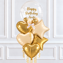 A birthday balloon bouquet with a large clear round balloon reading 'Happy Birthday Aoife' in gold script, surrounded by metallic gold and champagne-colored foil balloons in heart, star, and round shapes, tied with a white ribbon, set against a white paneled wall.