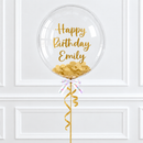 A clear round birthday balloon with gold foil lettering that reads 'Happy Birthday Emily' and gold confetti inside, tied with a white ribbon and gold curling ribbon against a white paneled wall.