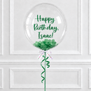 A large clear round balloon with green script text reading 'Happy Birthday, Isaac!' and green confetti inside, tied with a white ribbon and green curling ribbon against a white paneled wall.