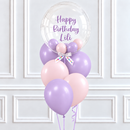 A balloon bouquet with a large clear round balloon reading 'Happy Birthday Lili' in purple script, surrounded by pastel purple, pink, and peach latex balloons tied together with a white ribbon, set against a white paneled wall.