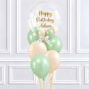 A balloon bouquet decoration featuring a large clear round balloon with gold 'Happy Birthday Adam' script, surrounded by pastel green, cream, and peach latex balloons tied together with a white ribbon, set against a white paneled wall.