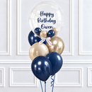 A birthday balloon bouquet with a large clear round balloon reading 'Happy Birthday Owen' and several metallic navy, gold, and navy balloons tied together with a white ribbon, set against a white paneled wall.