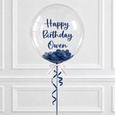 A clear round birthday balloon with navy blue script text reading 'Happy Birthday Owen' and navy blue confetti inside, tied with a ribbon and hanging against a white paneled wall.