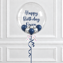 A clear round birthday balloon with the message 'Happy Birthday Owen' in navy script, floating above smaller navy and silver balloons tied with a patterned ribbon and navy curling ribbon, set against a white paneled wall.