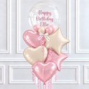 A birthday balloon bouquet with a large clear round balloon reading 'Happy Birthday Ellie' in pink script, surrounded by metallic pink and champagne star and heart foil balloons tied together with a pink ribbon, set against a white paneled wall.