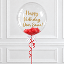 A clear round balloon with gold foil text reading 'Happy Birthday Dear Emma!' and red heart confetti near the bottom, tied with a white bow and red ribbon against a white paneled wall.