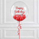 A clear round balloon filled with red script text reading 'Happy Birthday Samantha' and topped with several red balloons, tied with a white ribbon and red curling ribbon, set against a white paneled wall.